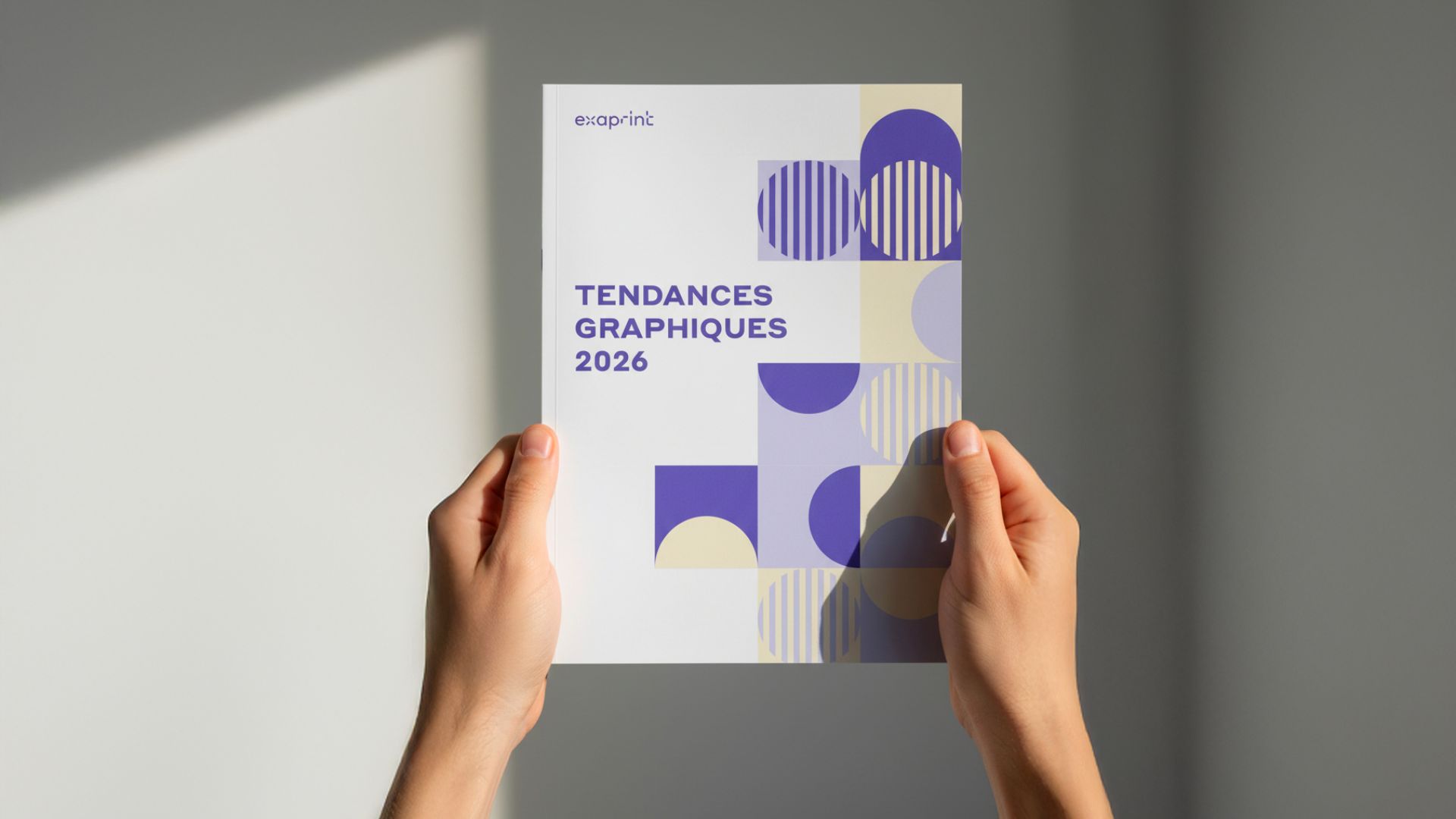 Gros plan sur la première de couverture du Guide des Tendances Graphiques 2026 tenu entre les mains d'un professionnel. Le visuel met en avant une composition graphique de formes géométriques et un nuancier de violets profonds, symbole de l'innovation et de l'expertise print d'Exaprint.